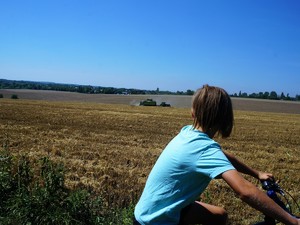 chłopiec siedzi na rowerze, znajduje się na drodze przy polu, po którym jedzie kombajn oraz traktor