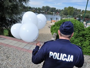 policjant stoi tyłem, w ręce trzyma pęk balonów, stoi przed schodami prowadzącymi na plażę zbiornika wodnego