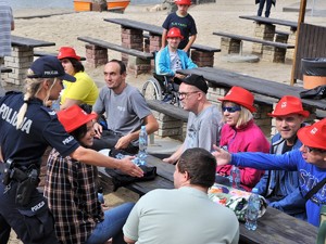 policjantka podaje dłoń mężczyźnie siedzącym wraz z innymi przy stoliku na plaży przy zbiorniku wodnym