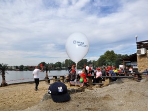 na piasku leży policyjna czapka, obok znajduje się balon z logo wodzisławskiej Policji, który jest wbity w piasek, w tle widać osoby siedzące na ławkach przy plaży zbiornika wodnego