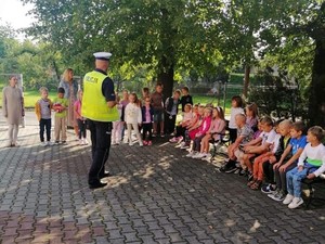 policjant stoi przed siedzącymi przed nim dziećmi, prowadzi spotkanie profilaktyczne