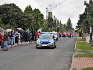 ulicą jedzie policyjny radiowóz, za nim maszerują uczestnicy dożynkowego korowodu