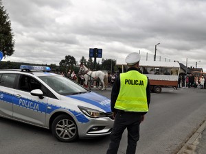 obok radiowozu stoi policjant, zabezpiecza przejazd dożynkowego korowodu