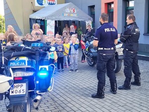 policjanci stoją przed budynkiem obok policyjnych motocykli, przed nimi stoją przedszkolaki i ich opiekunki