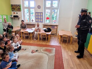 przed siedzącymi na podłodze przedszkolakami stoi policjant, prowadzi spotkanie profilaktyczne