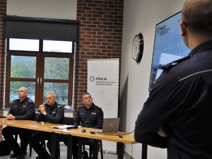 trzej policjanci siedzą przy stole, obok nich stoi inny policjant, który patrzy na wyświetlacz powieszony na ścianie