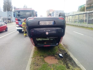 pomiędzy pasami jezdni, na pasie zieleni na dachu leży pojazd, obok pojazdu stoi strażak