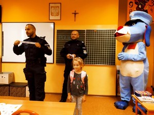 w sali lekcyjnej, policjant stoi obok uczennicy. Pokazuje odblaskową opaskę, za nimi stoi drugi policjant i maskotka śląskiej Policji, Pies Sznupek