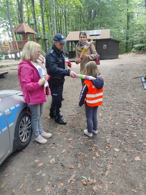 na terenie leśnym policjant pasuje na starszaka dziecko, dotyka specjalną kredką jego ramienia. obok niego stoi nauczycielka