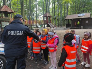 policjant stoi tyłem, przekazuje grupie dzieci stojącej przed nim odblask