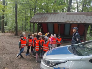 przed grupą dzieci stoi policjant, rozmawia z nimi, jedno z dzieci ma podniesioną do góry rękę