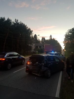 na pierwszym planie znajduje się uszkodzony pojazd, który stoi na jezdni, przed nim stoi policyjny radiowóz, który ma włączone sygnały świetlne, pojazdy te są wyprzedzane przez inne auta