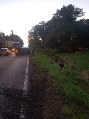 na zdjęciu widać stojący na jezdni uszkodzony pojazd, w tle w zaroślach widać drugi pojazd, który ma włączone światła