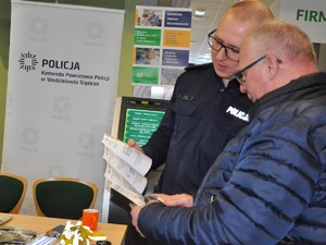 policjant w mundurze stoi obok mężczyzny, pokazuje mu ulotki profilaktyczne