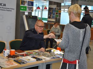 przed policjantem siedzącym przy stoliku stoi kobieta, która w ręku trzyma ulotkę profilaktyczną