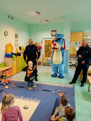 policjanci stoją w przedszkolnej sali obok Sznupka, maskotki policyjnej. Przed nimi stoi dziewczynka ubrana w policyjną kamizelkę, przodem do nich siedzą na podłodze przedszkolaki