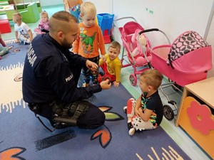 policjant kuca przed siedzącymi na podłodze przedszkolakami, pokazuje kajdanki, uśmiecha się