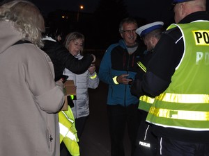 na zdjęciu dwaj policjanci stoją przed mężczyzną, obok którego stoi kobieta, przed nią dwie kolejne kobiety, urzędniczki Starostwa Powiatowego. Trwa akcja profilaktyczna podczas której piesi otrzymują elementy odblaskowe