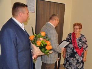 na zdjęciu widać jak komendant powiatowy składa życzenia kobiecie, za nim stoi zastępca komendanta powiatowego, który trzyma bukiet kwiatów