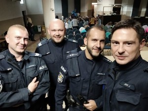 policjanci pozują do wspólnego zdjęcia, w tle widać uczniów, którzy przygotowują się do udziału w spotkaniu profilaktycznym