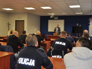 nieumundurowany policjant stoi przed uczestnikami szkolenia, siedzącymi przed nim na krzesłach