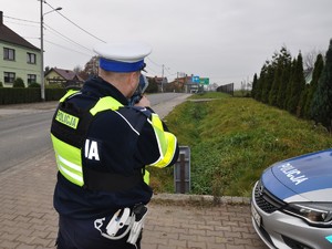 policjant mierzy prędkość przejeżdżających pojazdów, stoi przy drodze