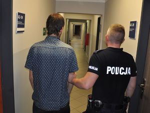 policjant podtrzymuje zatrzymanego za ramię. Mężczyźni stoją do zdjęcia tyłem