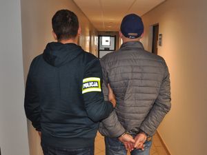 nieumundurowany policjant przytrzymuje zatrzymanego mężczyznę, który ma na rękach założone kajdanki. Mężczyźni stoją na korytarzu tyłem do zdjęcia