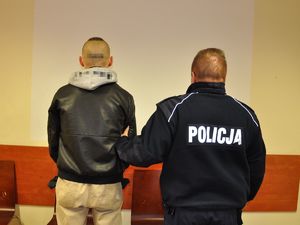 policjant stoi obok zatrzymanego mężczyzny, podtrzymuje go za ramię, mężczyźni stoją tyłem