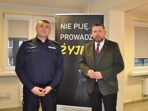 Zastępca Komendanta Powiatowego Policji w Wodzisławiu Śląskim stoi obok prezydenta Miasta Wodzisław Śląski, pozują do wspólnego zdjęcia na tle bilbordu z hasłem kampanii profilaktycznej