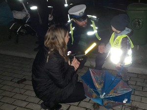 na zdjęciu widać policjanta, który kuca obok dziecka i z nim rozmawia, sytuacji przygląda się kobieta, która również kuca blisko nich