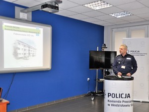 Komendant stoi przed siedzącymi podczas narady policjantami, przemawia do nich