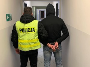 policjant ubrany w kamizelkę z napisem policja stoi obok mężczyzny, który ma na rękach założone kajdanki