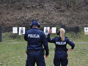 Policjant stoi obok policjantki, która trzyma jedną rękę w górze, drugą sięga po broń