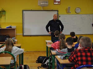 w sali lekcyjnej policjant prowadzi spotkanie profilaktyczne dla uczniów, którzy siedzą w ławkach