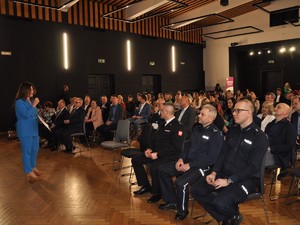 na sali na krzesłach siedzą zaproszeni goście, policjanci, strażak oraz inne osoby ubrane na galowo