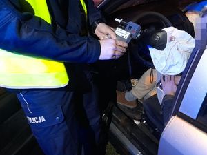 na zdjęciu widać rozbity samochód, w nim mężczyznę i stojącego obok policjanta, który kieruje w stronę kierowcy alkomat