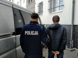 na zdjęciu widać stojącego obok radiowozu policjanta, który trzyma za rękę zatrzymanego mężczyznę , stoją tyłem do zjęcia