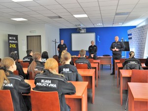3 policjanci stoją przed uczniami klas mundurowych siedzących w ławkach