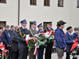 podczas uroczystości stoją obok siebie przedstawiciele służb mundurowych, trzymają w rękach  bukiety kwiatów z symbolicznymi wstążkami w barwach narodowych