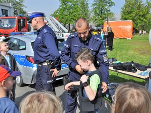 policjant zakłada chłopcu kamizelkę kuloodporną, pozostałe dzieci przyglądają się temu