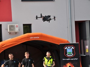 trzej strażacy stoją przed namiotem profilaktycznym, jeden kieruje dronem