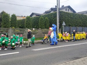 Zdjęcie przedstawia maskotkę oraz grupy dzieci idących w parach.