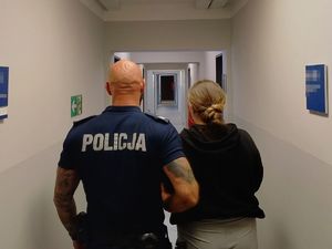 Policjant stoi obok zatrzymanej kobiety. Stoją tyłem do zdjęcia.