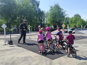 na zdjęciu widać policjanta który stoi obok grupy dzieci na rowerach, wszyscy znajdują się na terenie miasteczka ruchu drogowego