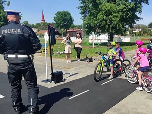 na zdjęciu widać policjanta który stoi obok grupy dzieci na rowerach, wszyscy znajdują się na terenie miasteczka ruchu drogowego