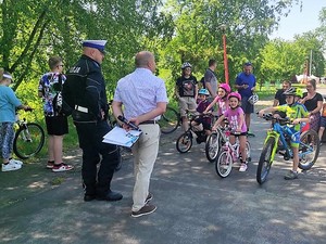 na zdjęciu widać policjanta który stoi obok grupy dzieci na rowerach, wszyscy znajdują się na terenie miasteczka ruchu drogowego, policjant rozmawia z mężczyznom