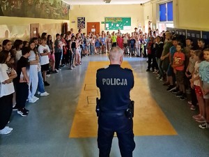 na szkolnym korytarzu widać zebranych uczniów oraz policjanta, który prowadzi z nimi pogadankę