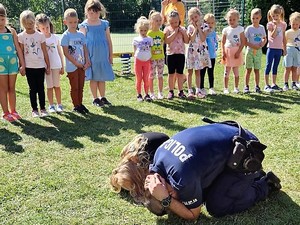 przed stojącymi w szeregu dziećmi, dziewczynka oraz policjant przyjmują pozycję żółwia