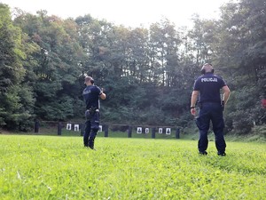 policjant strzela z broni długiej do tarczy, obok niego stoi policyjny instruktor strzelań